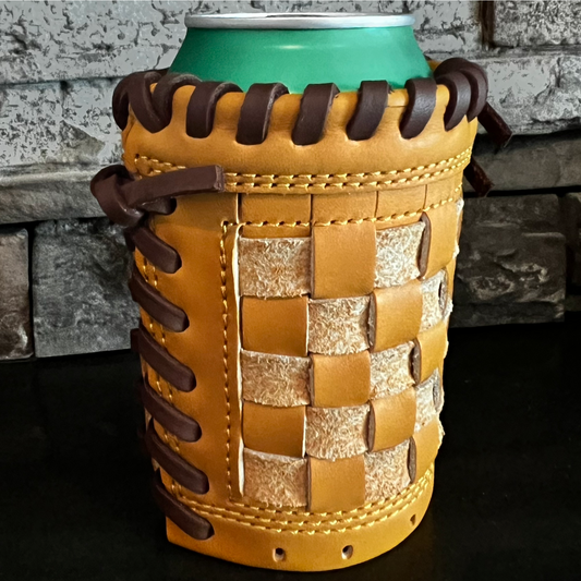 A tan leather can cooler with dark brown braided laces and a woven leather front panel, shown holding a green can against a dark tabletop and stone wall background.